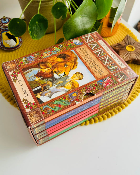 A boxed set of children's books titled 'NARNA' with illustrated covers, arranged on a yellow pom-pom placemat on a white table with potted plants and decorative items in the background.