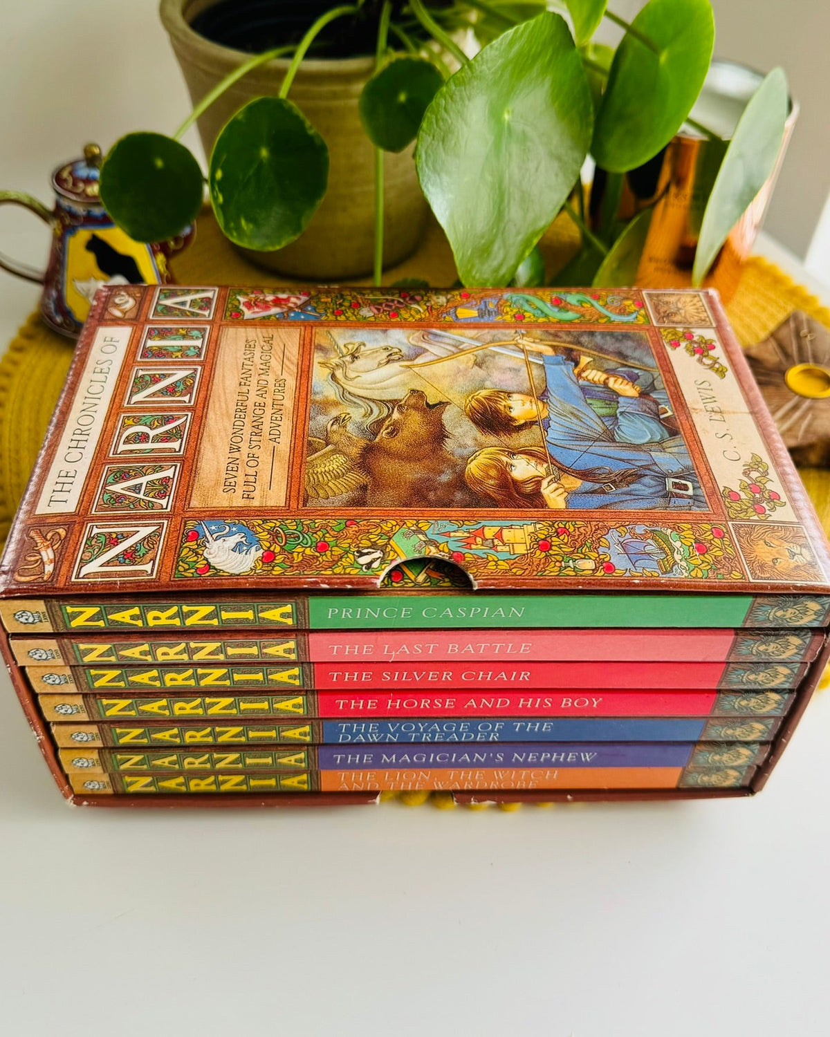 A boxed set of The Chronicles of Narnia books displayed on a yellow woven mat, with a potted green plant and small decorative objects in the background.