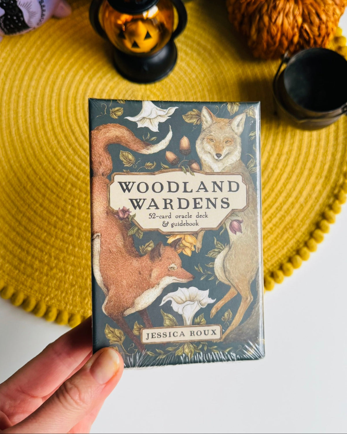 A hand holds a boxed card deck titled "Woodland Wardens" by Jessica Roux, featuring illustrated woodland animals on the front. The box is photographed over a yellow woven placemat with decorative objects in the background.