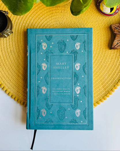 A teal hardcover book titled 'Mary Shelley: Frankenstein' lies on a yellow woven placemat, surrounded by decorative props including a green plant, a black cup, a small ceramic dish, and a wooden star-shaped candle holder on a white surface.