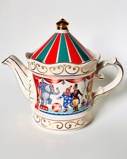 A vintage porcelain teapot with a red and green striped lid, gold trim, and a colorful circus-themed illustration on the body, set against a plain light gray background.