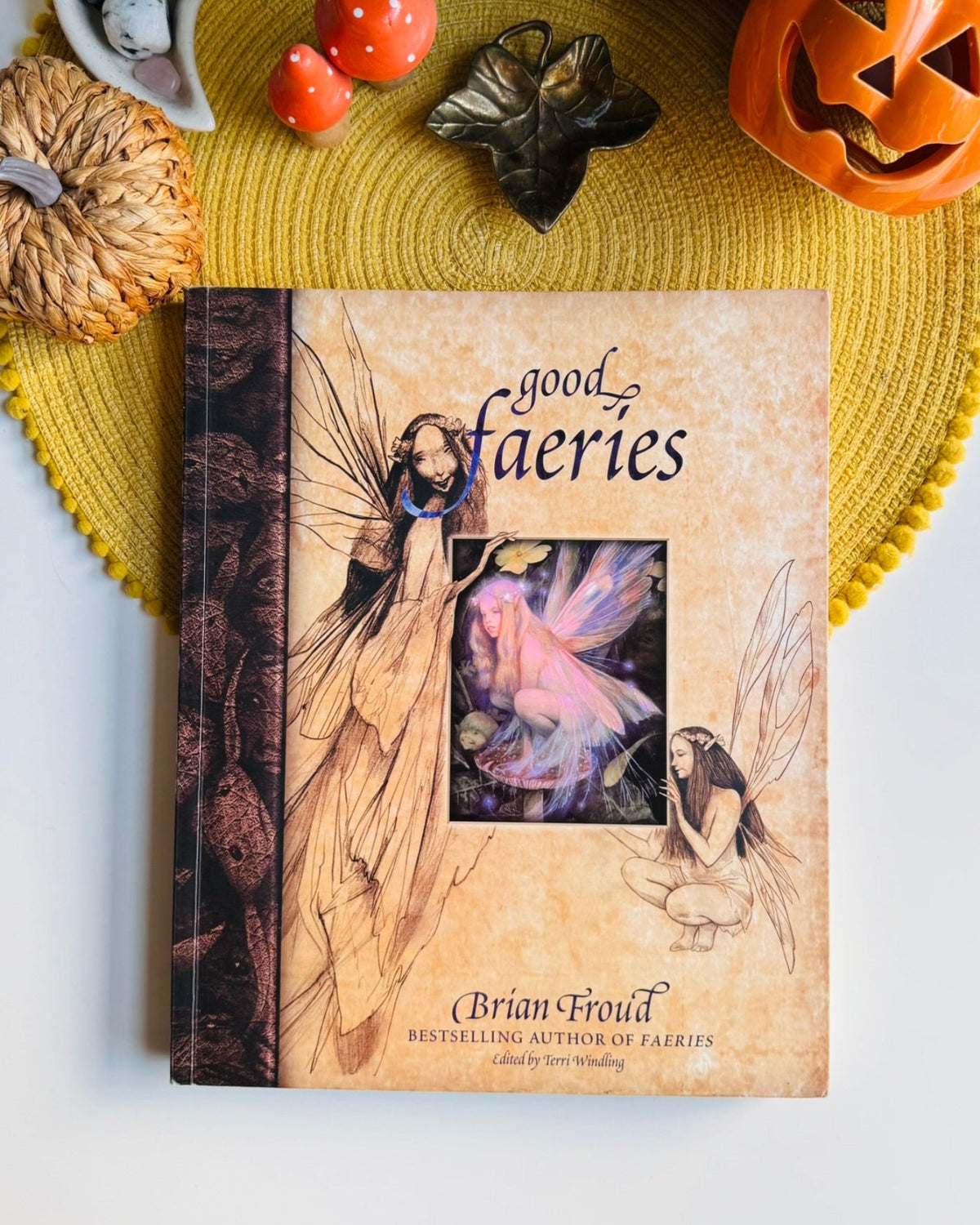 A book titled "good fairies" by Brian Froud lies on a yellow woven placemat, surrounded by Halloween-themed props including a carved pumpkin, small pumpkins, a leaf, and decorative objects on a white surface.