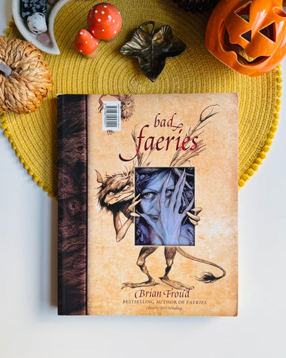 A book titled "bad fairies" by Brian Proud is displayed on a yellow woven placemat with Halloween-themed props including a carved pumpkin, small pumpkins, and dried leaves on a white surface.