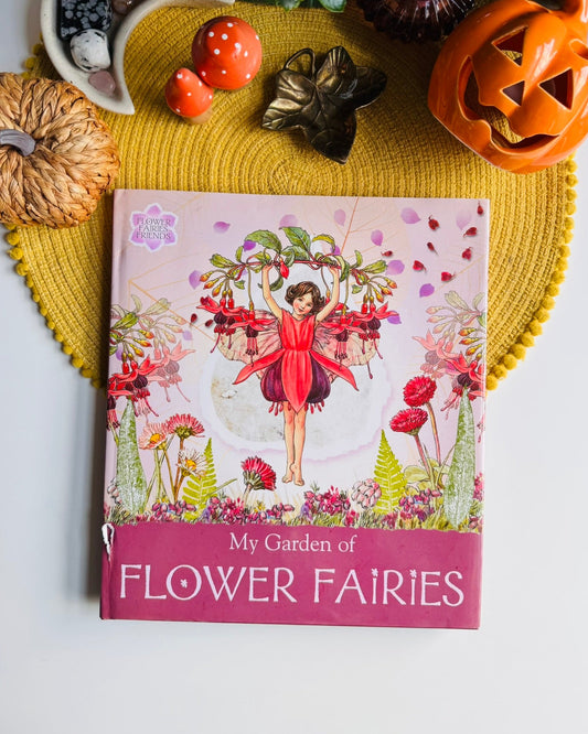 A children's book titled 'My Garden of Flower Fairies' is displayed on a yellow woven placemat with autumn-themed props including a carved pumpkin, small gourds, and a plant.