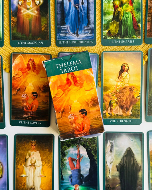 A Thelema Tarot deck with its box placed on top of several illustrated cards, arranged on a white surface with a gold rope border. The front of the box reads 'THELEMA TAROT' and shows a fiery orange-toned illustration.