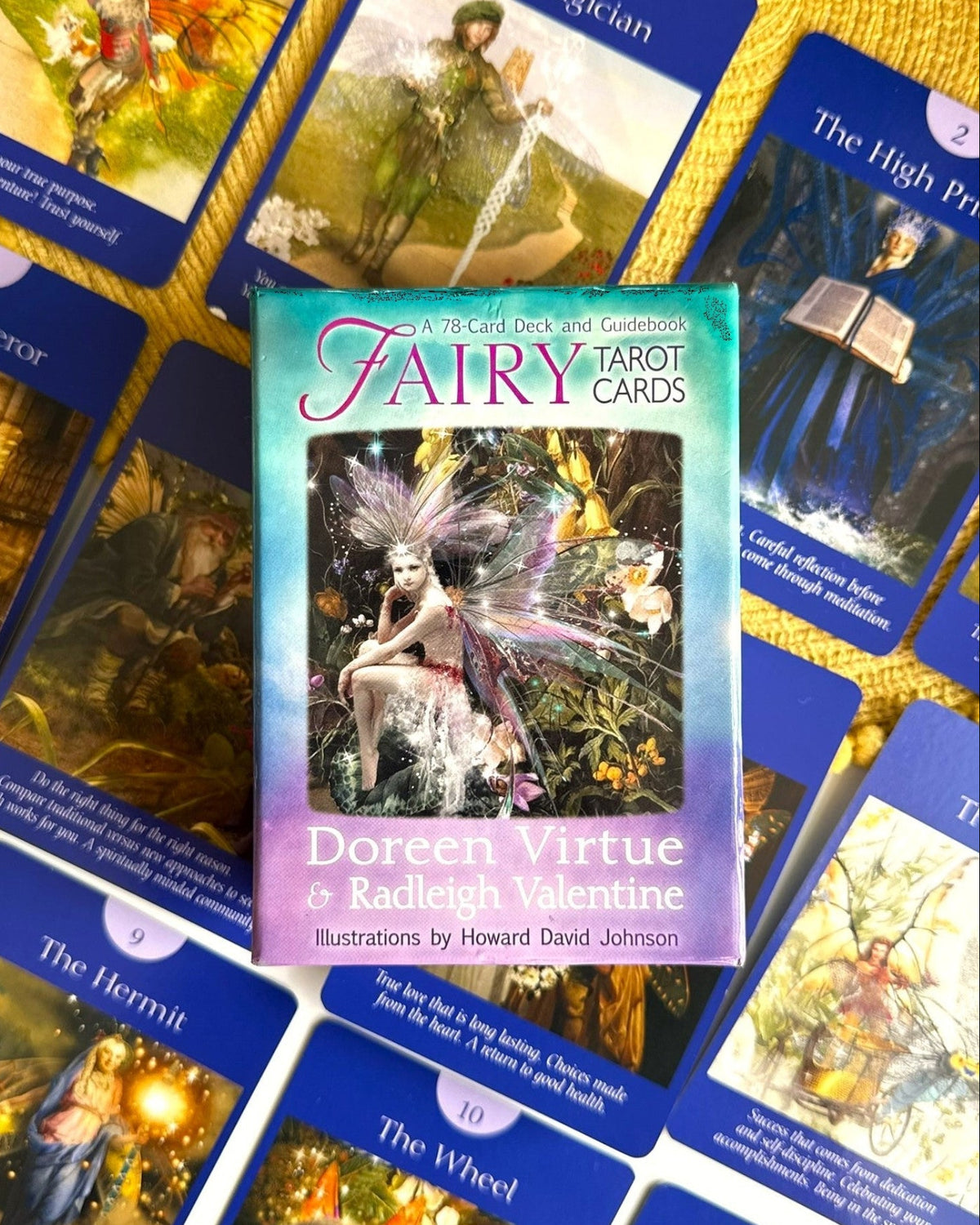 A boxed deck of fairy tarot cards is centered on top of several illustrated tarot cards spread out on a yellow textured surface. The front box reads 'Fairy Tarot Cards' and 'Doreen Virtue & Radleigh Valentine.