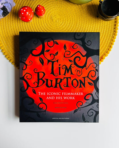 A book titled 'Tim Burton: The Iconic Filmmaker and His Work' by Tim Burton is displayed in a styled flat-lay on a white surface, surrounded by a yellow woven placemat, decorative red apples, and small cups.