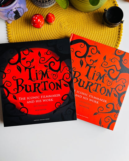 Two Tim Burton book covers are arranged on a white surface with a yellow woven placemat and small decorative props in the background. One cover is black with a large orange circular design and the other is orange with black swirling tree-like patterns.