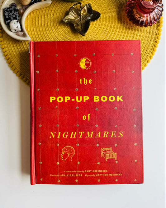 A red hardcover book titled "the POP-UP BOOK of NIGHTMARES" with yellow lettering, photographed from above on a white surface with a yellow woven placemat and decorative objects around it.
