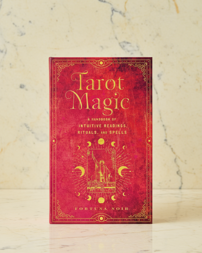 A tarot card deck box titled 'Tarot Magic' by Loretta Noir, standing upright on a light marble surface against a beige marble background. The box is deep red with gold ornate border details and gold lettering.